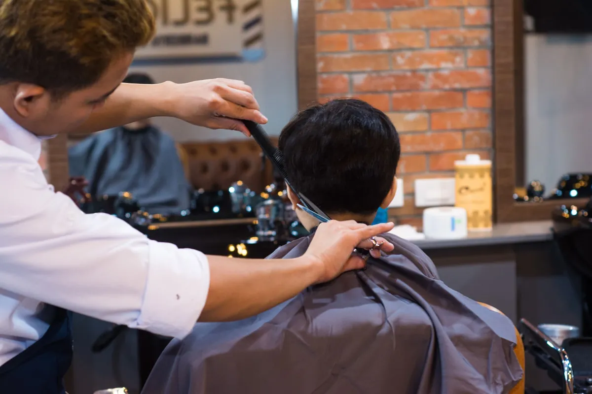 A barber combing and cutting a client's hair in a brick-walled shop.