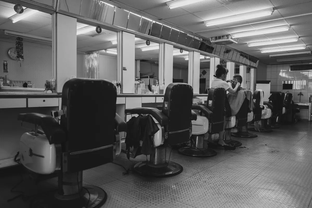 Black-and-white view down a row of classic barber chairs in a busy shop, one barber mid-cut.