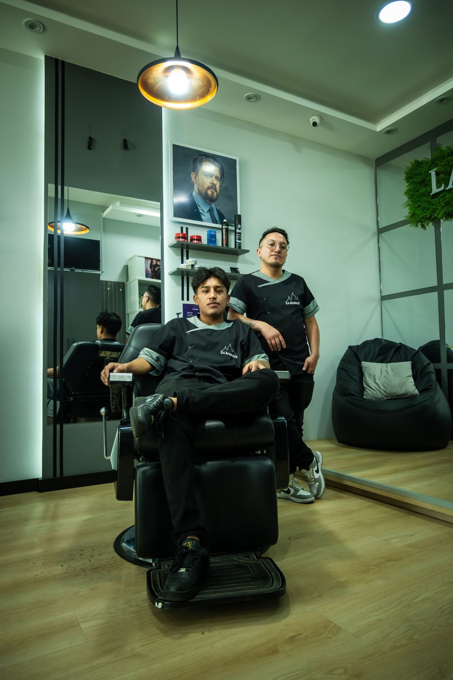 Two barbers posing in their shop — both sides of the chair, in one place.