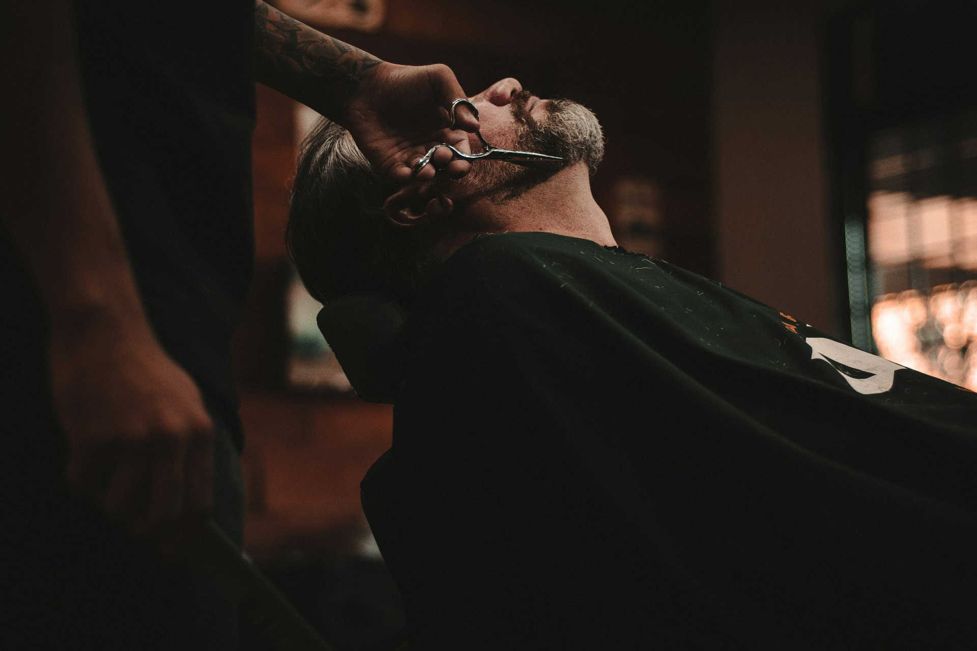 A barber trimming a client's beard with scissors in a warmly lit shop — the everyday math behind barber chair cost.
