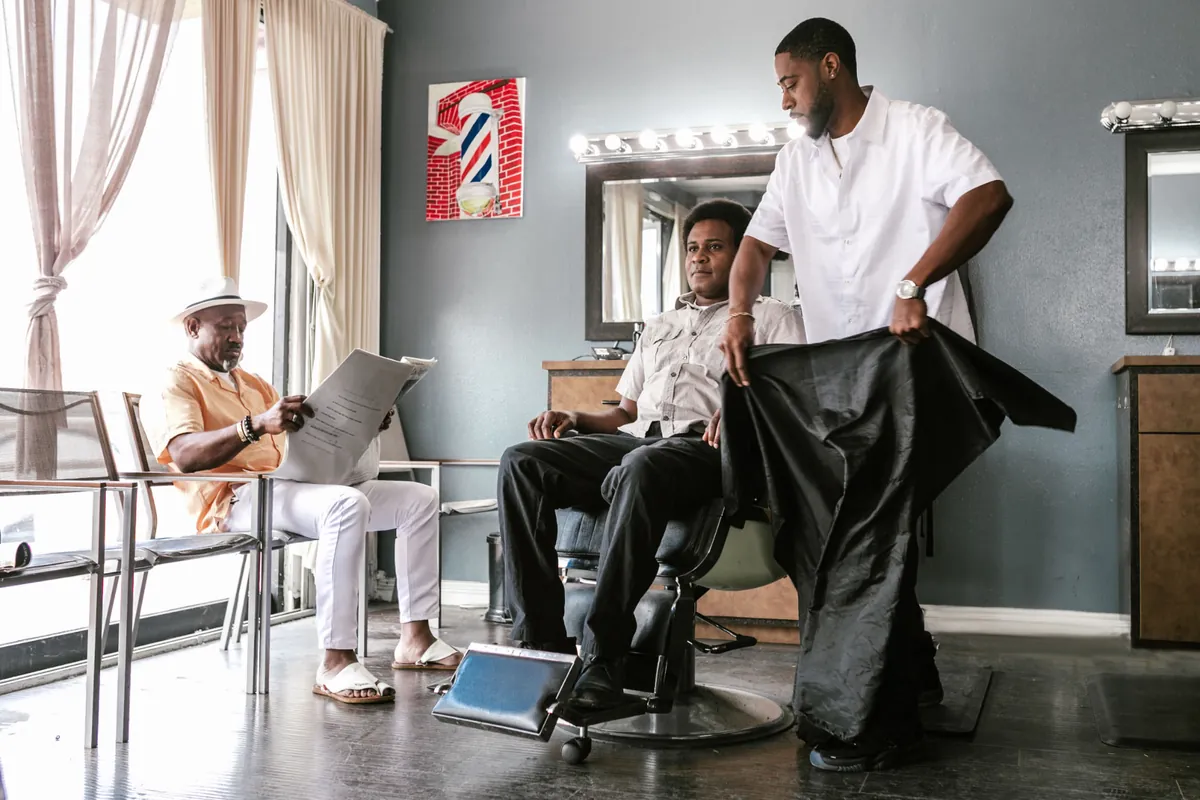 A barber draping a cape over a seated client in a bright neighborhood barbershop.